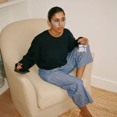 A woman sitting on a chair in the living room wearing under-eye patches drinking a glass of water