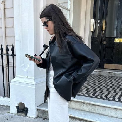British style influencer Anna Howard poses on a London street looking at her phone wearing a black leather jacket, gray T-shirt, and white jeans