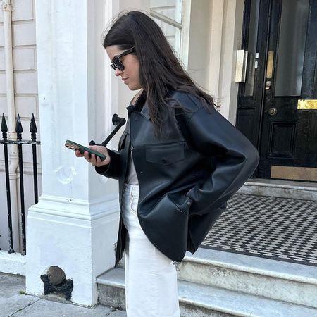 British style influencer Anna Howard poses on a London street looking at her phone wearing a black leather jacket, gray T-shirt, and white jeans