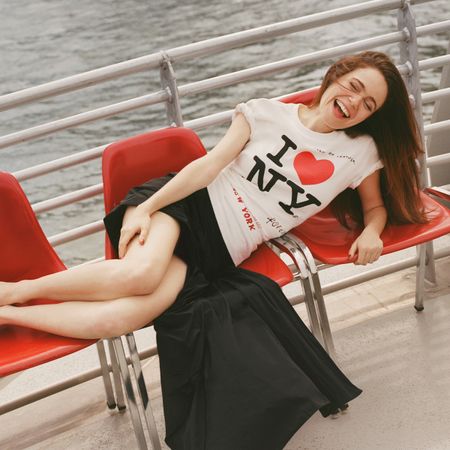 Photo of Molly Gordon laughing sitting across three red chairs outside on a ferry boat. She is wearing a 'I heart NY' t-shirt with a black skirt. 