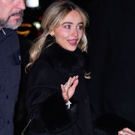 Sabrina Carpenter wears white satin platform heels with a longline black coat and a black leather handbag.