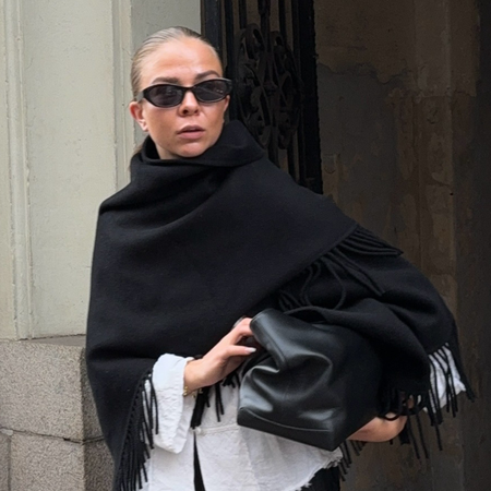 Scandi woman wearing black fringe scarf coat with black leather bag, black sunglasses, and white button down.